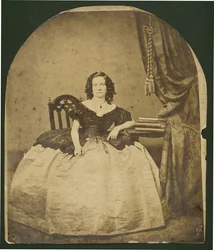 Ganzkörperporträt einer sitzenden, lockigen Frau in einem Kleid mit weitem Rock